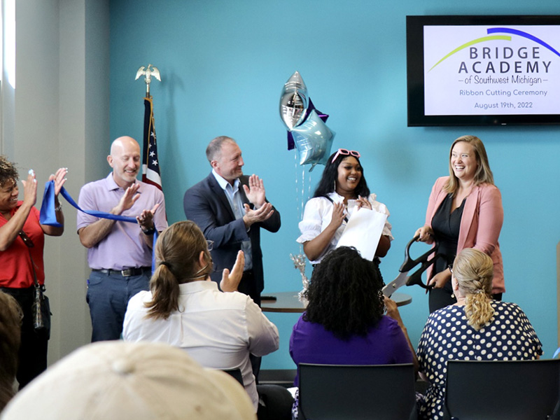 Bridge Academy of Southwest Michigan's administrator smiling after cutting the ribbon at their ribbon cutting ceremony. Board members, the Center's executive director, and a student are smiling and clapping.