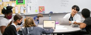 Six students from Renaissance Public School Academy working together at a table. Some are working on laptops, one is reading, and two students are talking to each other.