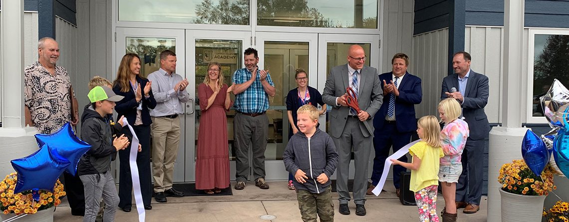 Copper Island Academy ribbon cutting ceremony. Staff and students gathered at front door clapping, holding ribbon that has been cut, and a student running.
