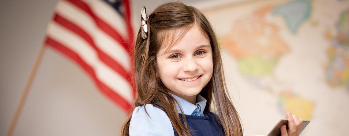 Student from Ivywood Classical Academy smiling for the camera, standing in front of the U.S.A flag, and holding a tablet.