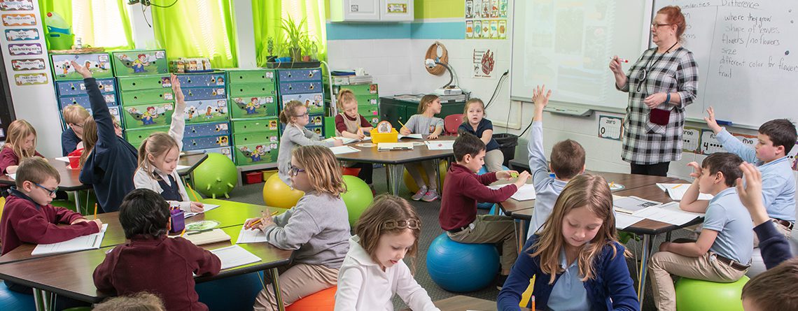 Teacher standing in front of classroom of students at Holly Academy. The students are sitting on balance balls at their tables. Some students are raising their hands and some students are writing.
